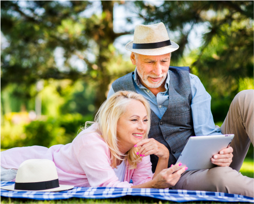 A middle-aged couple laying on a blanket outside looking at a tablet